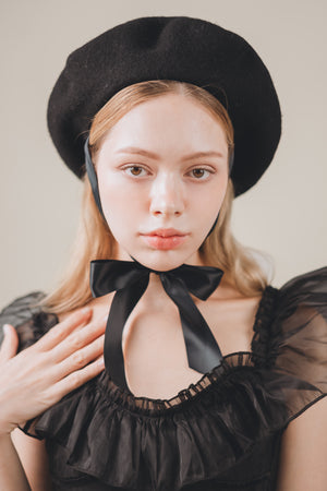 Woman wearing a black beret and black dress with a bow neckline against a neutral background