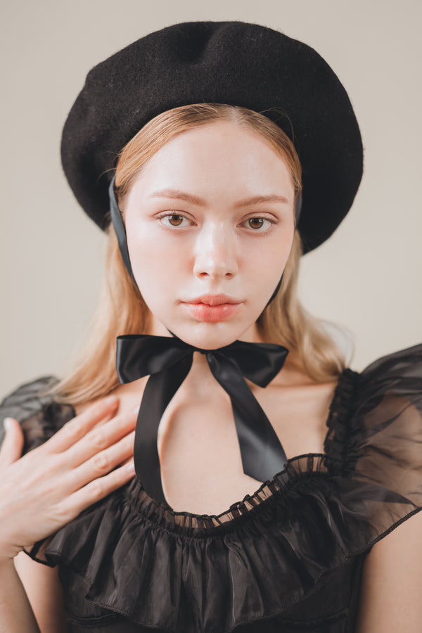 Woman wearing a black beret and black dress with a bow neckline against a neutral background