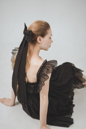 Woman in a black dress with sheer sleeves and a large bow sitting on a light gray background