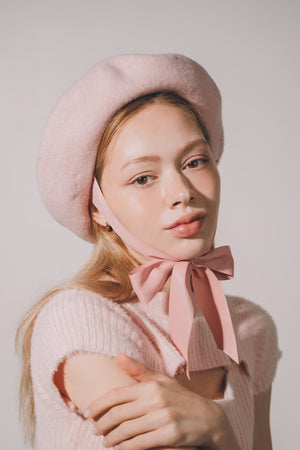 Woman wearing a pink beret and sweater with a bow, against a plain background